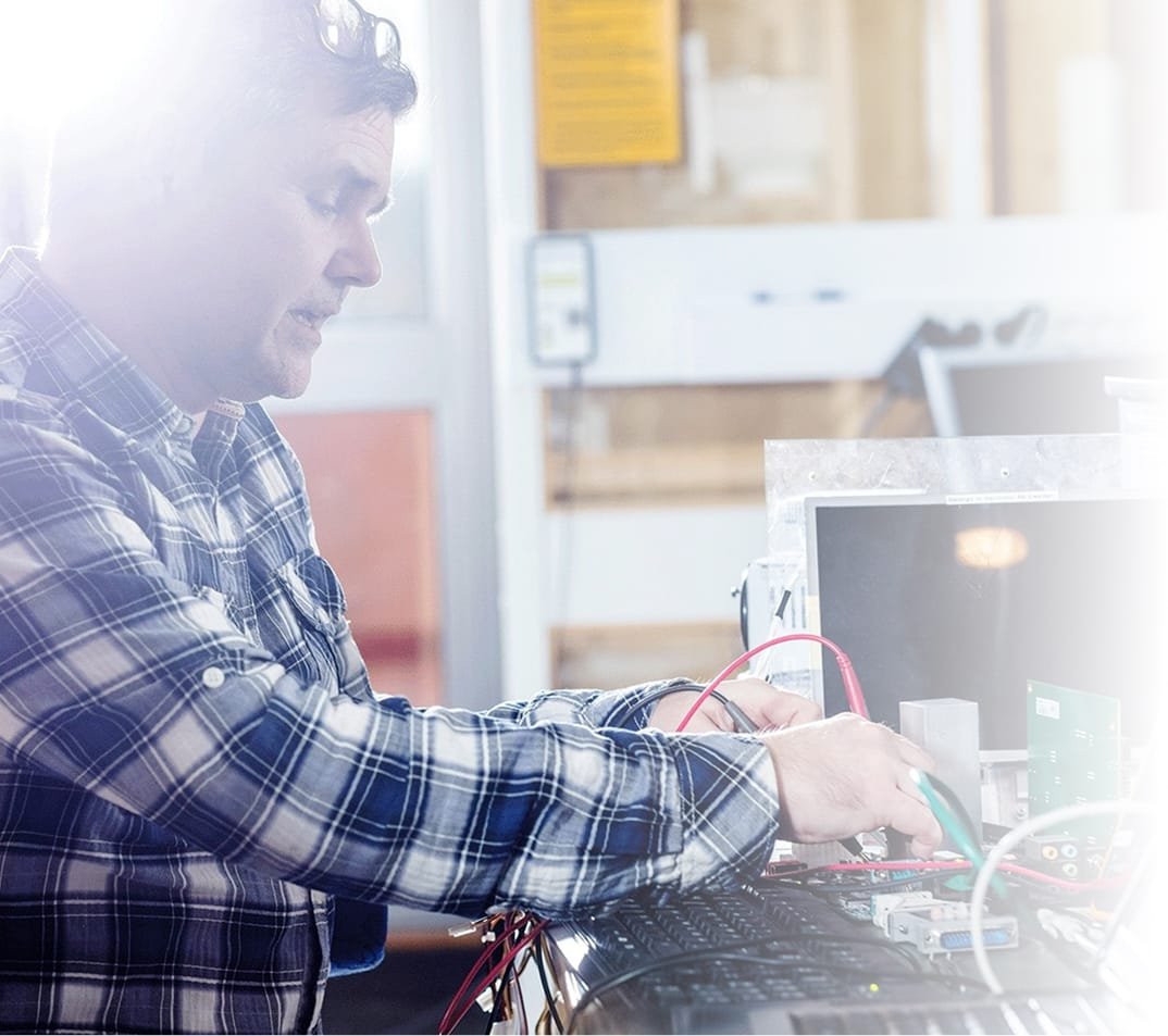 Medium shot of a technician in a plaid shirt working at an electronics bench: holding red and green leads attached to a PCB and modules beside a keyboard. Workshop background is out of focus with strong backlight coming from the left.